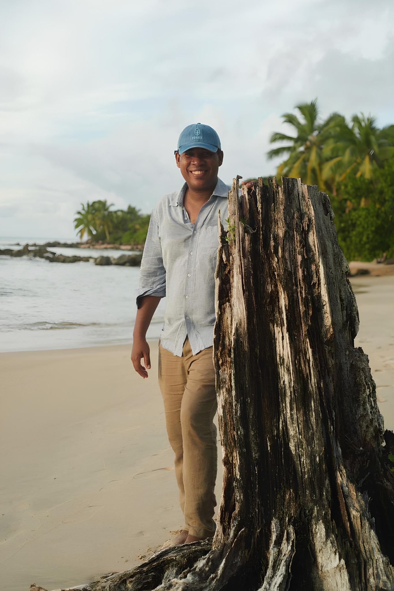 Jimmy Mahasolo, Resort Manager at Voaara Madagascar, smiling beside a weathered tree stump on a palm-lined beach.